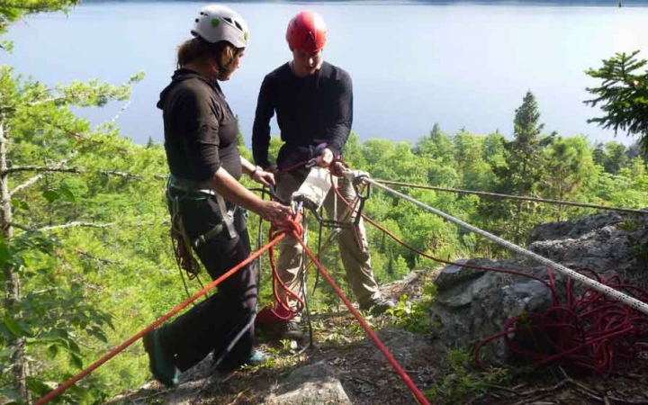 two people wearing rock climbing gear are attached to ropes at the top of a cliff while one gives instruction to the other
